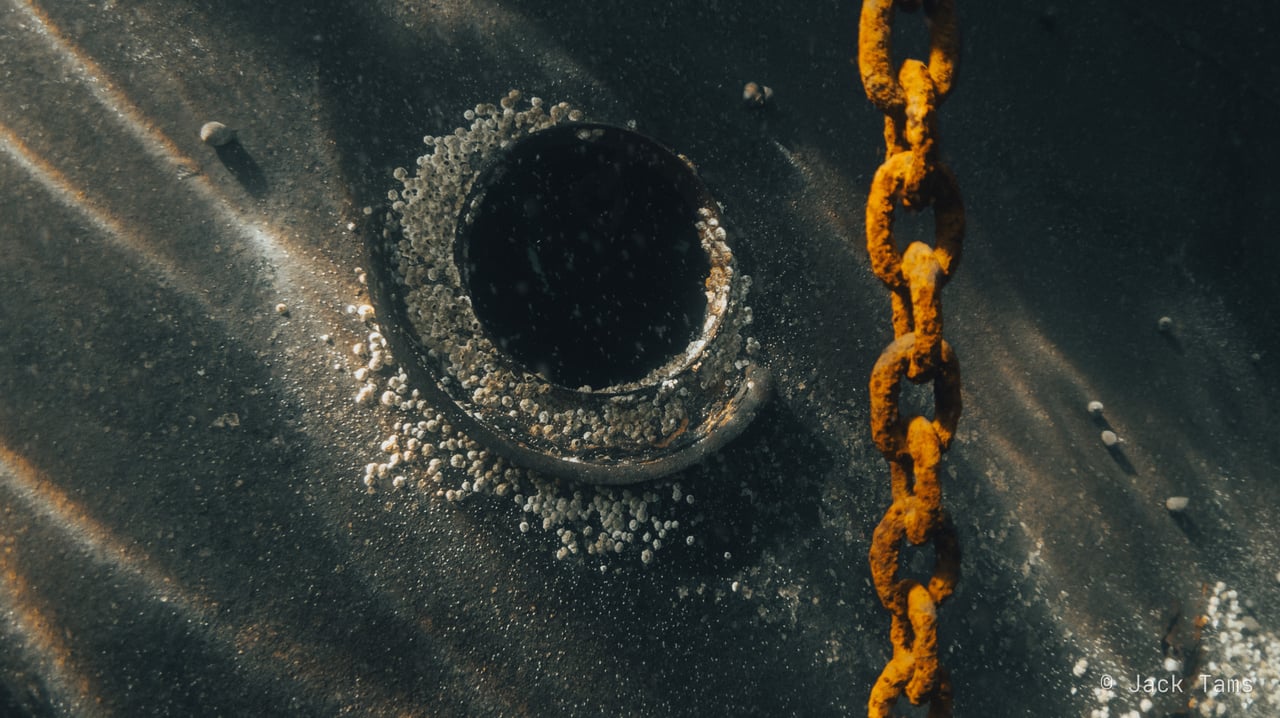 A porthole on the hull plating in the shallows, a rusted chain hanging alongside.